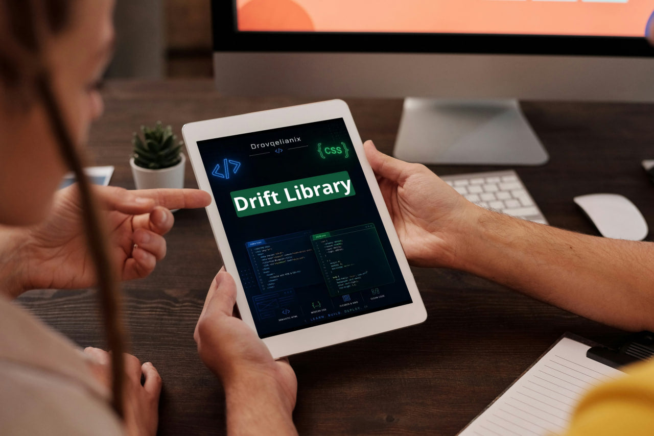 A person holds a tablet showing a digital coding interface labeled 'Drift Library' while another person points at the screen in a workspace setting with a computer, keyboard, and small plant in the background.